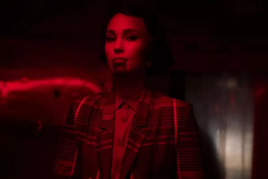 Kaneza Schaal in a dimly lit room bathed in intense red light, wearing a patterned jacket. The dramatic lighting and shadows create a cinematic atmosphere.