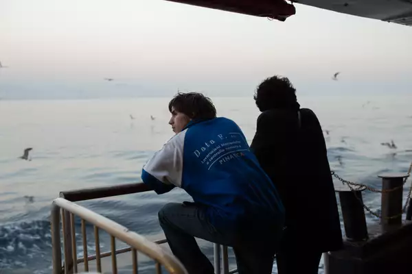 Still from Crossing. Two characters with their backs turned, stood by railings looking out at sea.