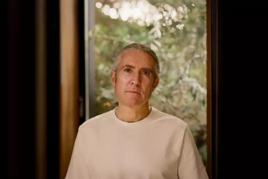A portrait of Richard Russell wearing a light-coloured shirt, standing in front of a window with a blurred green outdoor background.