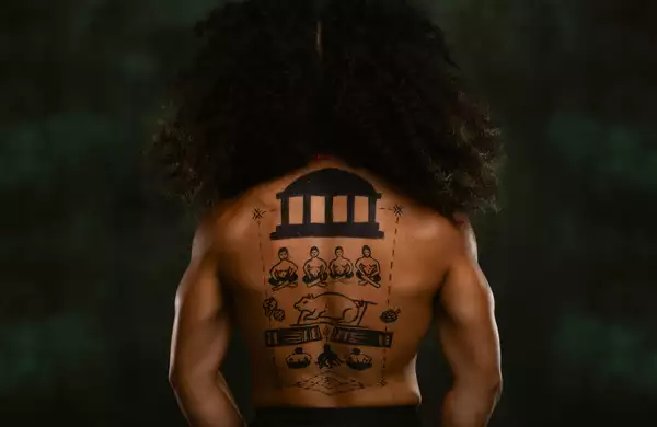 A member of FAFSWAG with their back turned away from the camera. They are topless with hand-drawings on their back. The background is dark green.