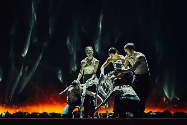 A still from Figures in Extinction [1.0] showing performers gathering around and holding a skeletal structure on a dimly lit stage with fiery hues in the background.