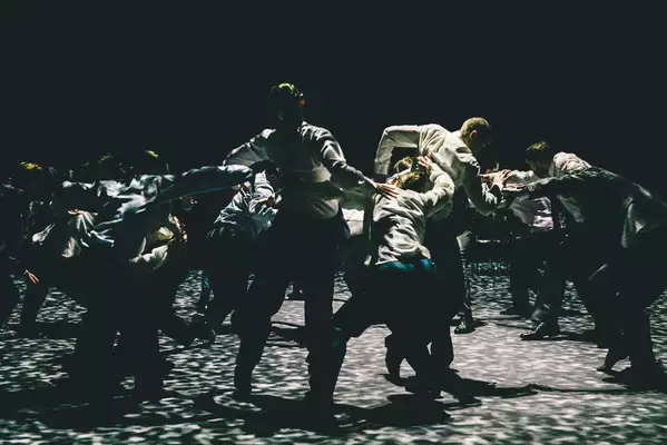 A still from Figures in Extinction [2.0] showing performers on stage wearing suits, making large contorted shapes with their bodies under spotted lighting