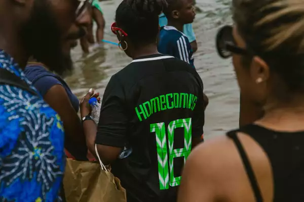 An image of people standing in the sea in Lagos, Nigeria, during Homecoming festival 2018. Most people are seen from the back or side; two people in the foreground frame the image, and in the centre a person wears a football shirt with 'Homecoming 18' printed on the back, red sunglasses and gold hoop earrings.