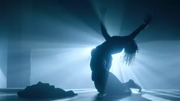 A photograph of a dancer on their knees, toes curled and arms outstretched. They can only be seen in silhouette as a bright spotlight provides a strong backlight.