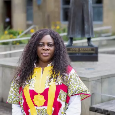 Headshot of Chantelle Tindall wearing a red and yellow patterned top. They are stood outside in front of a statue of the suffragette Annie Kenney.