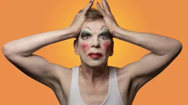 David Hoyle bold makeup and hands either side of his forehead against an orange background