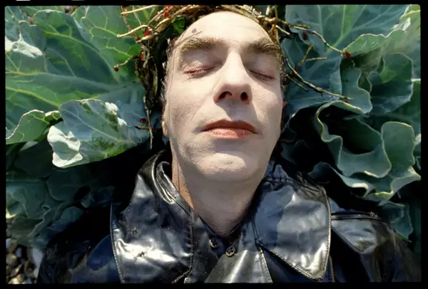 Still from Derek Jarman's The Garden (1990), showing a close up of Jarman's face. He is lying on a cabbage plant, wearing a black leather jacket and golden crown.