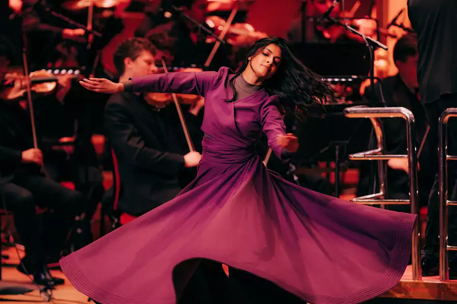 A dancer in a flowing purple dress gracefully spins on stage. Behind her, an orchestra dressed in black plays classical instruments.
