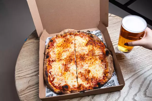 An open pizza box on a wooden table with a cheese pizza cut into four slices. A hand with red-painted nails holds a pint of beer beside it.