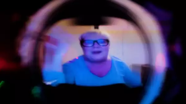 A white middle-aged woman with short blond hair and black rimmed glasses waves into a camera aperture. The image is distorted and out of focus.