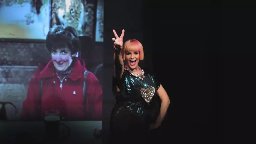 Kate O’Donnell making a peace sign, stood infront of a screen showing Hayley Cropper from Coronation Street