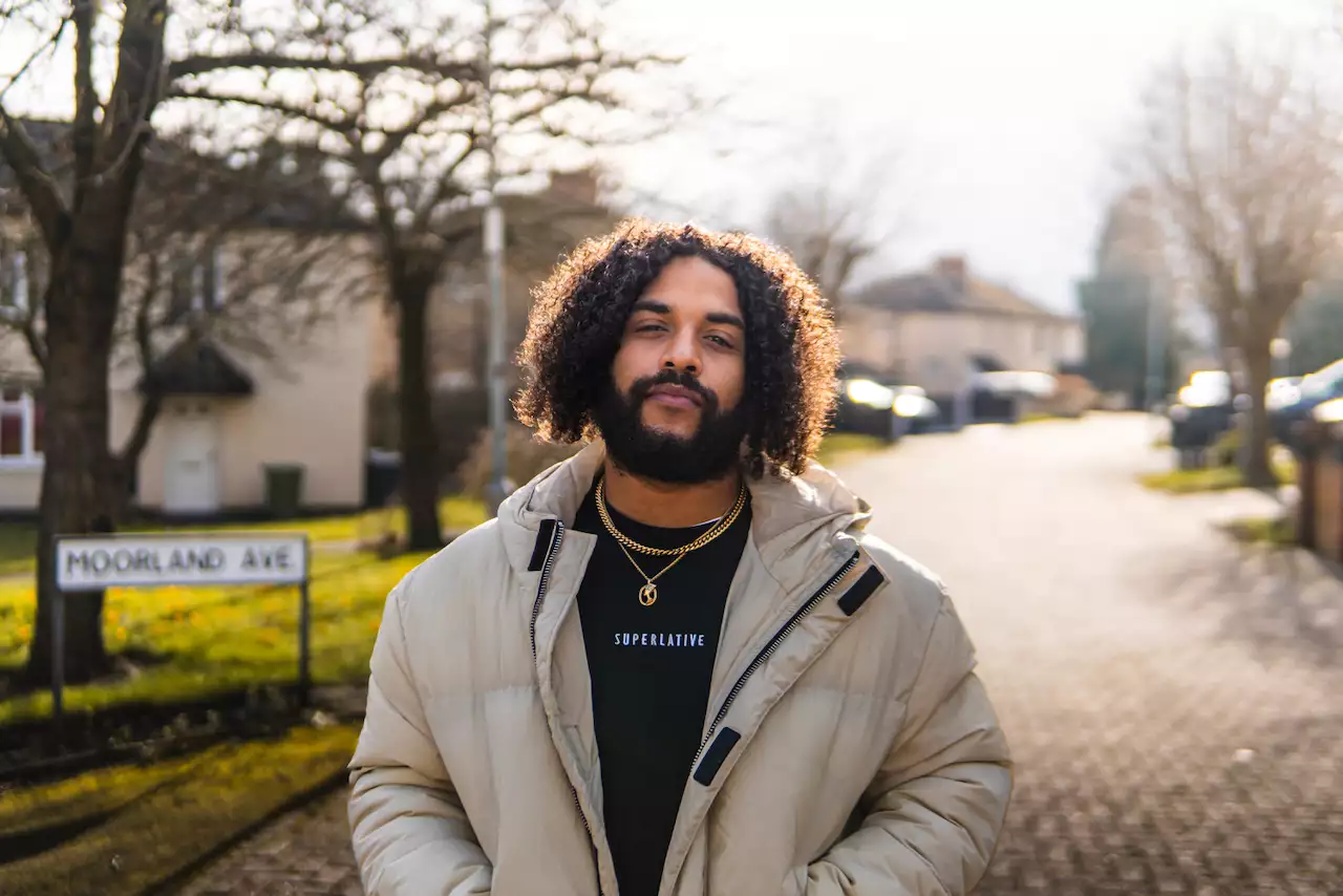 Corey Brown stands on a quiet street with bare trees and houses in the background, wearing a beige puffer jacket over a black shirt that says "SUPERLATIVE."