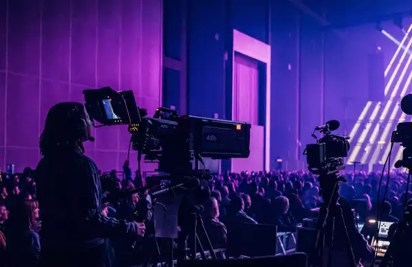 A camera operator films a live event in the Warehouse, which is filled with people. There is dramatic purple lighting with multiple cameras and bright stage beams.
