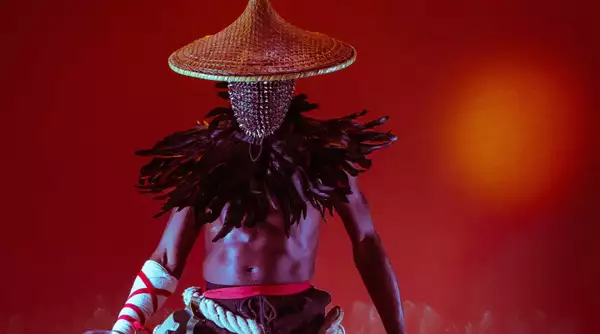 A photograph by Benji Reid, showing a man wearing an elaborate feathered headdress, which hides his face. He is standing against a deep red background.