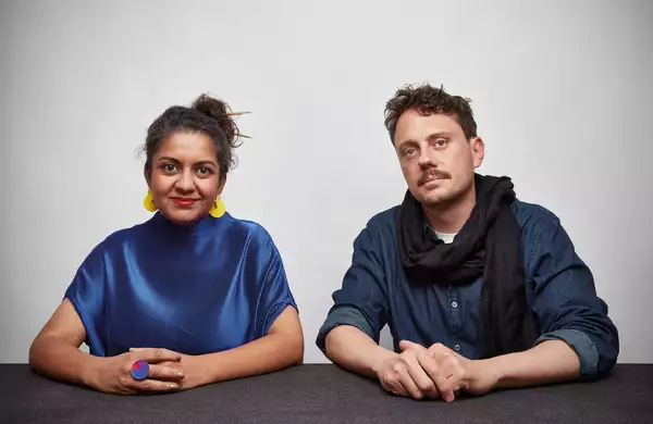 Two members of Superflux sat at a table, looking at the camera. The person on the left is wearing a shiny blue top and yellow earrings. The person on the right is wearing a blue shirt and black scarf.