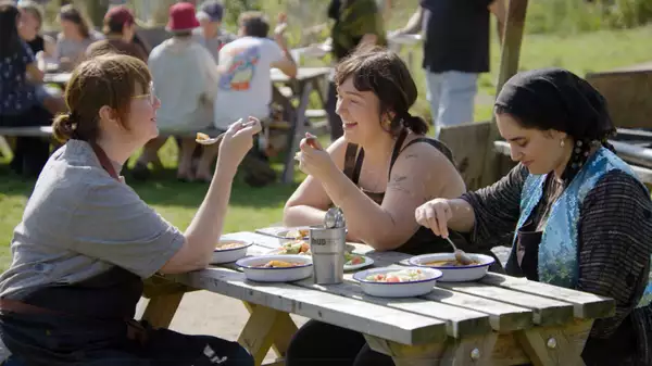Three people sat outside on a picnic table eating from bowls, talking and laughing.