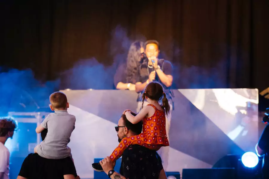 Two children sat on two adults' shoulders watching a performer on stage on Factory Square