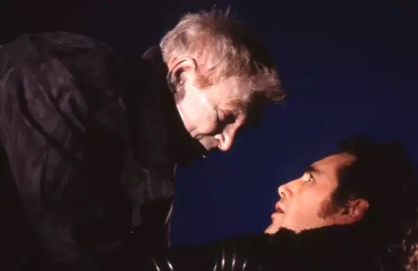 Russell Enoch towering over Mark Rylance, who is looking up at him in fear. Russell has his hands on Mark's neck. Both are wearing black coats.