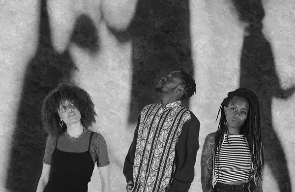 A black and white photograph of three members of The Untold Orchestra with shadowy figures in the background