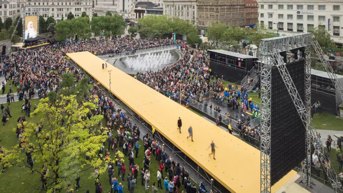 Three people walk down a large yellow catwalk, surrounded by a crowded audience in Piccadilly Gardens, Manchester