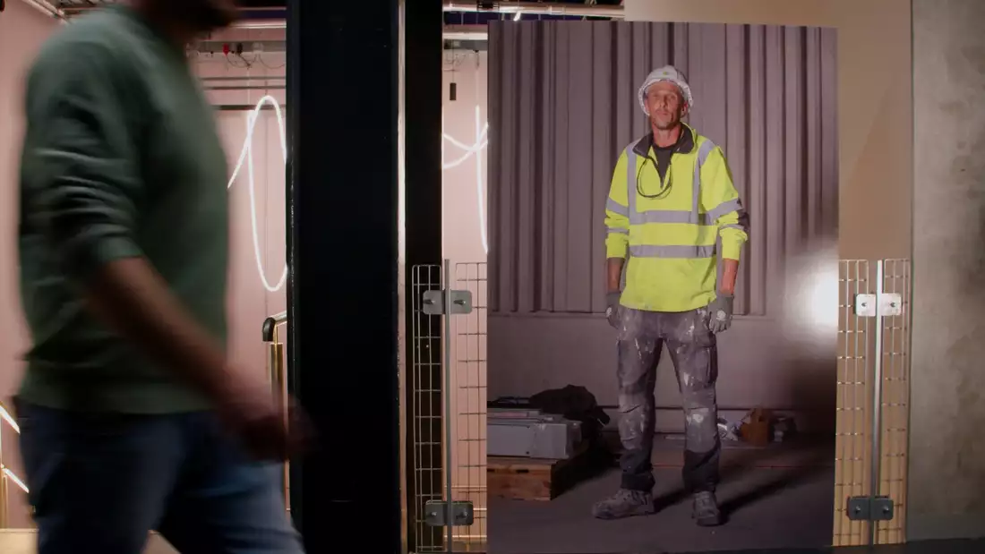 A person walking past a Ming de Nasty portrait of an Aviva Studios worker in a hardhat