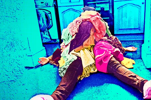 Woman lying on the floor of a kitchen with a pile oflaundry on top of her